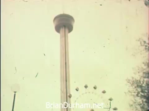 The still captures a view of the Tower of the Americas, a prominent feature at Hemisfair '68 in San Antonio. In the foreground, you can see elements of the fair, including a ferris wheel, which highlights the festive atmosphere of the event. The image reflects the juxtaposition of enjoyment and work, as secretaries participate in their professional duties amid the attractions designed for exploration and leisure.