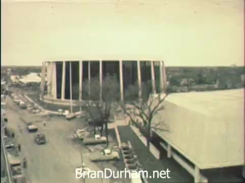 The still from 'News Item - Secretaries at Hemisfair '68' captures an aerial view of the Hemisfair venue in San Antonio. Prominent structures include the circular US Pavilion and a nearby building, framed by bare trees and a bustling street below. The scene reflects a blend of work and leisure, illustrating the dynamic atmosphere of Secretaries' Week amidst the backdrop of a major international expo.