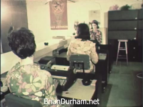 The still captures a busy office scene at Hemisfair '68 in San Antonio, showcasing women seated at desks, engaged in administrative tasks. They are dressed in vibrant floral patterns, suggesting a lively atmosphere during Secretaries' Week. The background features minimal decor, including a poster or banner, and shelves or filing cabinets, emphasizing their organized work environment amidst the excitement of the event. The juxtaposition of their diligent work against the festive context of Hemisfair highlights their commitment to office responsibilities.