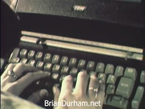 The still features a close-up of hands typing on an IBM typewriter, reflecting the busy atmosphere of Secretaries' Week at Hemisfair '68 in San Antonio. The scene emphasizes the dedication of secretarial staff as they manage correspondence amidst the allure of the event's attractions, like the US pavilion. The focus on the typing hands captures the blend of professional duties with the festive environment surrounding them.