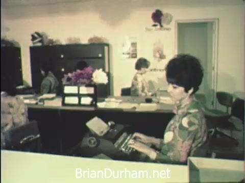 The still from 'News Item - Secretaries at Hemisfair '68' depicts a group of secretaries engaged in office work during Secretaries' Week at the San Antonio Hemisfair. The environment is busy yet vibrant, with one secretary in the foreground typing on a typewriter, adorned in a colorful floral blouse. Behind her, others are focused on their tasks amidst an organized workspace, featuring filing cabinets and decorative elements. The scene captures the balance between the demands of professional duties and the lively backdrop of the event.