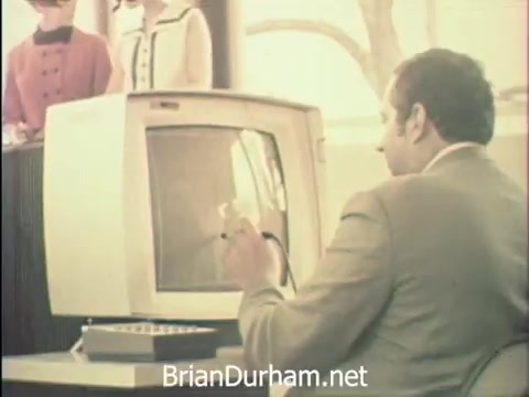 The still features a man in a suit seated in front of a monitor, likely from the late 1960s, as indicated by the style of the technology and clothing. He appears to be interacting with the computer, possibly typing or using a device, while a group of women in period attire stands in the background. The context of the image highlights the observation of Secretaries' Week at Hemisfair '68 in San Antonio, emphasizing the blend of office duties with the festive atmosphere of the exposition. The scene captures the intersection of professional responsibilities and the vibrant social environment of the world's fair.