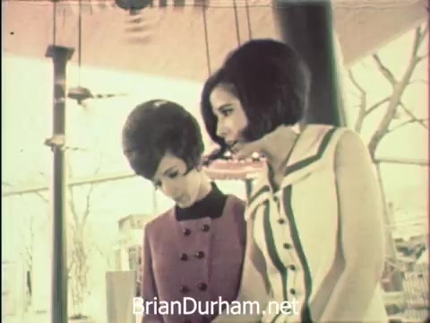 The still depicts two women dressed in stylish 1960s attire, engaged in a moment of focus at Hemisfair '68 in San Antonio during Secretaries' Week. The setting hints at a blend of professional duties and the vibrant atmosphere of the exposition, as they balance the demands of office work with the allure of the event. The background suggests elements of the fair, reinforcing the contrast between their responsibilities and the festive context surrounding them.