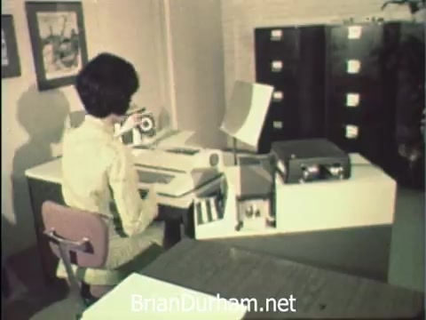 The still from 'News Item - Secretaries at Hemisfair '68' captures a young woman seated at a retro office desk, focused on her work. She is surrounded by typical office equipment of the era, including a typewriter and filing cabinets. The setting reflects a blend of professional duties and the lively ambiance of the Hemisfair event. The context emphasizes the balance between office responsibilities and the unique experiences available at the fair during Secretaries' Week.