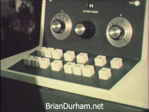 The still captures a close-up of a vintage control panel or device, featuring large knobs and a series of square buttons. This imagery reflects the technological vibe of Hemisfair '68, where secretaries are balancing office tasks with the vibrant atmosphere of the fair. The context highlights their role amid the festivities, maintaining correspondence while navigating the allure of various exhibits like the US pavilion.
