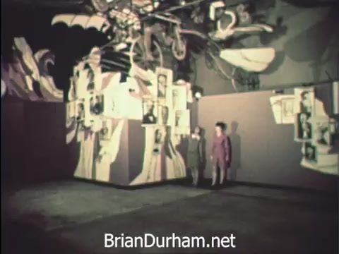 The still from 'News Item - Secretaries at Hemisfair '68' depicts two women standing in an artfully designed exhibition space, likely within the US pavilion at the Hemisfair. The backdrop features vibrant murals and photographs, creating a dynamic atmosphere. While appreciating the event, the women remain focused on their duties during Secretaries' Week, balancing the festive environment with their office responsibilities amidst the world's fair's intriguing offerings.