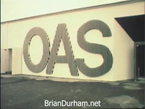 The still from 'News Item - Secretaries at Hemisfair '68' features the prominent lettering 'OAS' on a building, hinting at the Organization of American States. The backdrop reflects the setting of the Hemisfair in San Antonio, where secretaries are engaging in their professional duties during Secretaries' Week, balancing office responsibilities with the allure of the fair's attractions. The scene captures the essence of productivity amidst a vibrant environment.