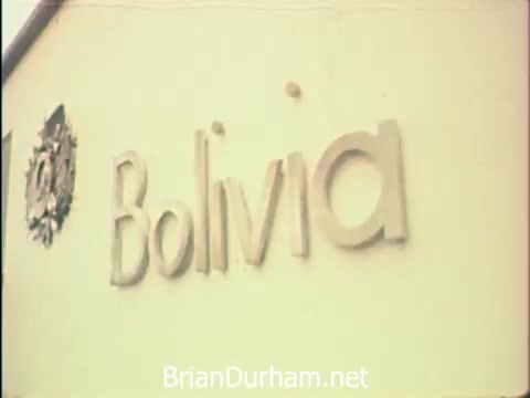 The still depicts a sign displaying the name 'Bolivia,' likely from the Hemisfair '68 event in San Antonio. The context suggests it was a busy week for secretaries, who managed office correspondence while also enjoying unique attractions at the fair, including the US pavilion. The image captures a moment that highlights both work and the cultural experience of the fair.