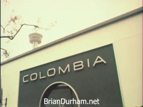 The still captures a building marked 'COLOMBIA' at the Hemisfair '68 in San Antonio, showcasing a moment during Secretaries' Week. It hints at the lively atmosphere of the event, with the iconic tower in the background, juxtaposing the vibrant experiences and professional duties of the secretaries amidst the world's fair setting. The focus on correspondence highlights their commitment, even while enjoying the diverse offerings around them.