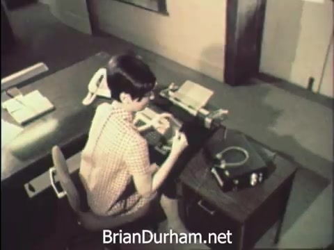 The still captures a young woman seated at a desk, engaged in office work during 'Secretaries' Week' at Hemisfair '68 in San Antonio. She is focused on typing or managing correspondence, surrounded by office supplies and a rotary phone. The setting reflects a blend of work and the vibrant atmosphere of the World's Fair, highlighting the daily routine of office secretaries amidst the exciting backdrop of the event.