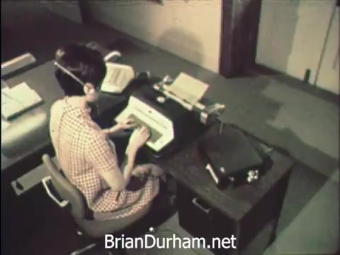 The still from 'News Item - Secretaries at Hemisfair '68' captures a moment in an office setting where a young woman in a checked dress is focused on typing at a typewriter. Her workspace includes papers, a binder, and an audio device, highlighting the busy atmosphere of handling correspondence. The context emphasizes the juxtaposition of office duties and the allure of the Hemisfair, showcasing the unique experiences of secretaries during Secretaries' Week.