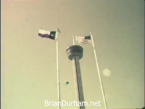 The still captures a sunny day at the Hemisfair '68 in San Antonio. Prominently featured are the flags of Texas and the United States, fluttering alongside a tall, cylindrical structure that signifies the Hemisfair's attractions, including the US pavilion. This image reflects the vibrant atmosphere of the event while hinting at the busy schedules of office workers observing Secretaries' Week, balancing their professional duties with the allure of the fair.