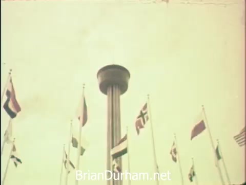 The still depicts a towering structure surrounded by various national flags, likely emphasizing the international spirit of the Hemisfair '68 event in San Antonio. The image captures a moment during Secretaries' Week, reflecting the blend of leisure and professional obligations faced by the working women's group as they navigate their duties amidst the fair's attractions. The overall tone suggests a celebration of their roles, with the fair serving as a backdrop for their experiences during this special week.