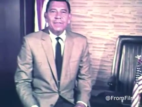 The still features Jack Webb, presenting a public service announcement for U.S. savings bonds and stamps from 1969. He is seated in a formal setting, wearing a light-colored suit and tie, with an American flag visible nearby. The background suggests a professional atmosphere, likely emphasizing the importance of saving and investing. The overall tone is serious and authoritative, typical of Webb's persona in public service campaigns.