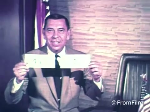 In this still from the 1969 PSA featuring Jack Webb, he is prominently displaying US savings bonds and stamps. He is dressed in a suit and tie, exuding a professional demeanor. Behind him, the backdrop includes a textured wall and the flags of the United States. The overall tone is focused on emphasizing the importance of saving and investing in U.S. savings bonds.