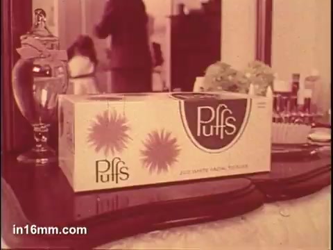 The still features a box of Puffs tissues prominently displayed on a wooden vanity or dresser. The packaging is colorful, showcasing the brand name 'Puffs' with stylized flower graphics. In the background, a figure is partially visible, indicating an indoor setting, likely a living room or bathroom, with personal items placed around the box, emphasizing a homey atmosphere. The overall tone is nostalgic and inviting, typical of vintage advertisements.