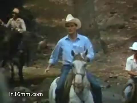 The still from the 1980 Peace Corps Public Service Announcement features a central figure riding a white horse, dressed in a cowboy hat and a light blue shirt. Surrounding him are other horse riders, with a backdrop of a wooded area. The image conveys a sense of adventure and teamwork, highlighting the rugged, communal spirit associated with the Peace Corps, underlined by the tagline, 'The toughest job you'll ever love.'
