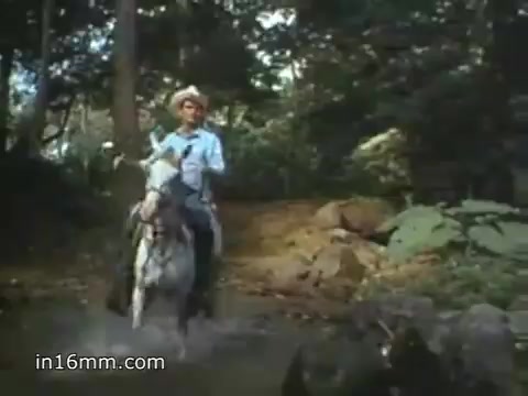 The still from the 1980 Peace Corps Public Service Announcement depicts a person on horseback crossing a shallow stream in a lush, green setting. The individual is wearing a light-colored shirt and a hat, embodying the adventurous spirit of the Peace Corps. This imagery aligns with the slogan 'The toughest job you'll ever love,' emphasizing the challenges and rewards of service in diverse environments. The vibrant natural backdrop highlights the connection to community and culture.