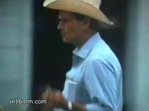 The still from the 1980 Peace Corps Public Service Announcement features a man in a wide-brimmed hat, dressed in a light blue shirt. He appears engaged in an activity, possibly in a rural or community setting. The overall tone reflects the themes of adventure and service, aligning with the slogan 'The toughest job you'll ever love.' The 16mm film grain adds a nostalgic quality to the image.