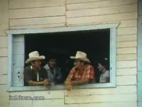 The still features a scene from the 1980 Peace Corps Public Service Announcement, showcasing a group of individuals wearing cowboy hats. They are gathered around a window of a rustic building, engaged in conversation. The atmosphere reflects camaraderie and community, highlighting the message of the Peace Corps about connection and service. The image has a vintage quality typical of 16mm film prints, evoking a sense of nostalgia.