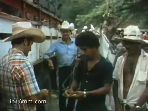 The still from the 1980 Peace Corps Public Service Announcement depicts a rural scene with people interacting. A man in a plaid shirt and cowboy hat is handing something to another individual, likely a local resident. In the background, two men observe, one wearing a light blue shirt and another sporting a white hat. The setting appears informal and community-focused, emphasizing connection and service in a tropical or subtropical environment, reflecting the announcement's theme of dedication to challenging but rewarding work.