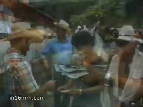 The still from the 1980 Peace Corps Public Service Announcement features a group of people engaging in a community activity. Individuals, including a man in a plaid shirt and a cowboy hat, are interacting with locals, demonstrating collaboration and friendship. The scene likely reflects the spirit of helping and cultural exchange, emphasizing the message: 'The toughest job you'll ever love.' The visual style suggests a documentary aesthetic typical of the era's public service announcements.