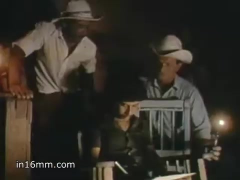 The still from the 1980 Peace Corps Public Service Announcement features three individuals in a dimly lit setting. Two men, wearing cowboy hats and casual shirts, lean over a seated woman, who appears to be focused on something in her lap. The atmosphere suggests a sense of collaboration and engagement, underscoring the message of the Peace Corps as a challenging yet rewarding experience. The tagline, 'The toughest job you'll ever love,' emphasizes the dedication required for service.