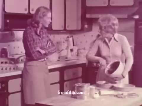 The still features two women in a kitchen, engaged in a lighthearted conversation. One woman stands with her arms crossed, watching as the other stirs a bowl, likely preparing a recipe. The kitchen has a vintage aesthetic typical of the 1970s, with wood cabinetry and classic cookware. The color tone is muted, characteristic of 16mm film prints from that era. The scene highlights a domestic setting focused on cooking and camaraderie.