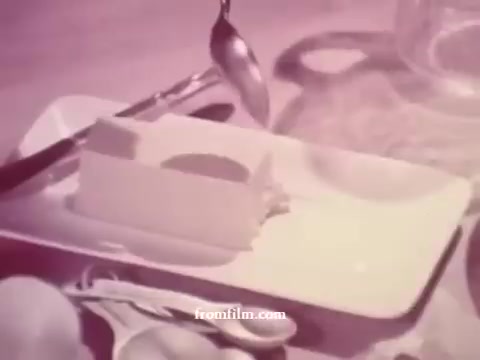 The still from 'Imperial Margarine: Spoon!' features a block of margarine on a rectangular plate, accompanied by a spoon, a knife, and several eggs. The scene is softly lit, with a pinkish hue, creating a warm and inviting atmosphere typical of 1970s advertising. The focus on the margarine suggests its usability in cooking or baking, emphasizing its appeal as a versatile kitchen staple.