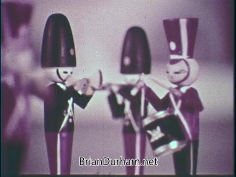 The still from the PSA 'Support Music In Your School' features a group of stylized toy soldiers or musicians playing instruments. They are depicted in a simplistic, colorful design, emphasizing their roles in a band. The background is softly blurred, directing focus on the figures, which likely represent the importance of music education in schools. The overall tone is playful and nostalgic.