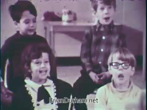The still from the PSA 'Support Music In Your School' features a group of four young children engaged in singing. They appear to be in a classroom setting, with the focus on their joyful expressions. Two boys are positioned in the back and two children, a girl and a boy, are in the front, with one child wearing glasses. The scene emphasizes the importance of music education in schools through the children’s enthusiastic participation.