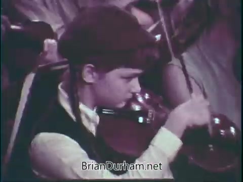 The still features a young violinist focused on playing in an orchestra setting. The child, dressed in period attire, showcases a dedication to music as they handle their instrument, emphasizing the importance of supporting music programs in schools. The image captures a moment of engagement and artistry in a performance.