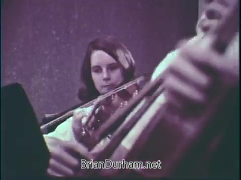 The still from the PSA 'Support Music In Your School' features a young student focused while playing an instrument, likely a violin. The image captures the dedication and engagement of youth in music education, emphasizing the importance of supporting music programs in schools. The background appears subdued, focusing attention on the student and their instrument.