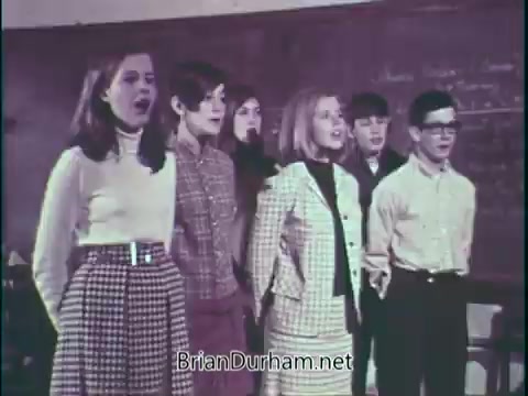 The still from 'Support Music In Your School - PSA' features a group of six students standing together, engaged in song. They appear to be in a classroom setting with a chalkboard in the background. The students are dressed in a mix of attire, including patterned skirts and button-up shirts, reflecting a vintage style. Their expressions convey enthusiasm and focus, emphasizing a message about the importance of music education in schools.