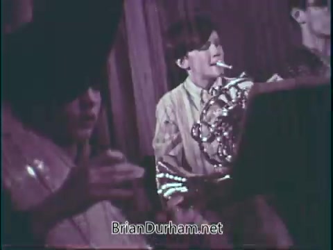 The still features a young musician playing a brass instrument, likely a French horn. He appears engaged and focused on his performance. Another individual partially visible in the foreground seems to be participating as well. The setting suggests a school or community music event, emphasizing the importance of music education. The overall tone conveys enthusiasm for supporting music programs in schools.