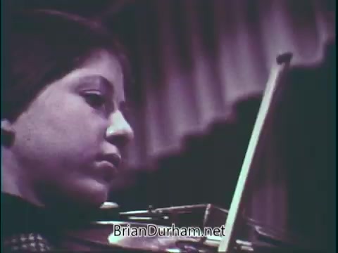 The still from 'Support Music In Your School - PSA' features a young violinist focused intently on playing. The close-up captures the profile of the child, emphasizing their concentration and dedication. The setting suggests a performance or practice environment, with a blurred background likely indicating an audience or a stage. The overall tone is serious and inspirational, underscoring the importance of music education.