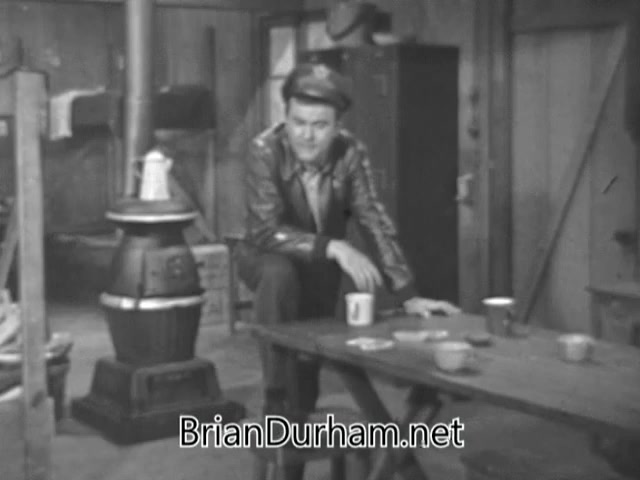 The 16mm still from the 'Bob Crane Army Air Guard PSA' features Bob Crane seated at a rustic wooden table in a military-style setting. He is dressed in a jacket and cap, conveying a casual yet authoritative presence. The background includes a stove and various items typical of a barracks environment. Crane's expression suggests a blend of seriousness and camaraderie as he addresses pilots leaving active service, reminding them about the benefits of flight pay while balancing the fun of making comedy with a sense of duty.