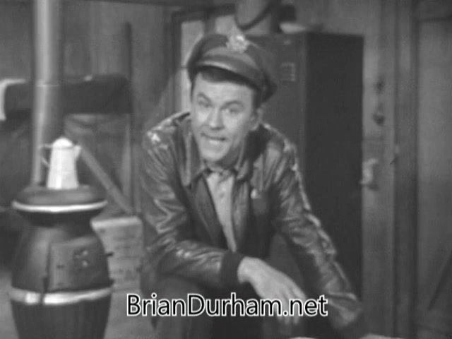 In this 16mm still from the 'Bob Crane Army Air Guard PSA', Bob Crane is depicted wearing a military-style jacket and cap, conveying a casual yet authoritative demeanor. He appears to be in a setting resembling a barracks, with a potbelly stove and wooden boxes around him. The black-and-white imagery enhances the nostalgic feel of the piece. Crane is engaging directly with the audience, likely discussing the importance of transitioning benefits for pilots leaving active service, blending humor with a serious message.
