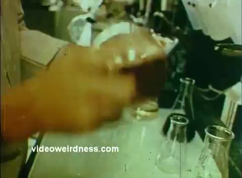 The still from the 'Sandoz Pharmaceuticals/Colors and Chemicals Commercial' features a close-up of a person's hand mixing or shaking a container. Surrounding the hand, laboratory equipment, including flasks and beakers, is visible, suggesting a scientific setting. The colors appear rich and somewhat muted, characteristic of 16mm film, conveying a vintage aesthetic. The context implies a focus on scientific processes, likely aimed at educating viewers about chemistry or pharmaceutical development.
