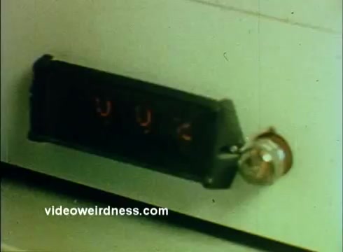The still from the 'Sandoz Pharmaceuticals/Colors and Chemicals Commercial' features a close-up of a digital display panel with illuminated numbers in a rectangular housing, likely indicating a measurement or count. The device appears to be part of a scientific instrument or laboratory equipment, enhancing the educational theme of the commercial aimed at classroom use. The overall aesthetic is characteristic of 16mm film, showcasing muted colors and a vintage quality.