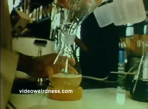 The still from the 'Sandoz Pharmaceuticals/Colors and Chemicals Commercial' features a laboratory setting where a person is pouring a liquid from a glass funnel into a round-bottom flask placed on a tabletop. The background includes shelves stocked with lab equipment and various containers, indicating a scientific environment. The image conveys a focus on chemical experimentation, likely aimed at promoting the responsibly of Sandoz Pharmaceuticals in contributing to educational science initiatives.