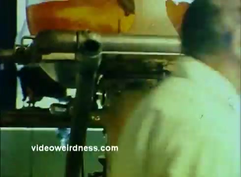 The still from the Sandoz Pharmaceuticals/Colors and Chemicals Commercial features an industrial setting, likely a laboratory or manufacturing area. In the foreground, a man in a lab coat appears to be in motion, possibly engaging with equipment. Behind him, machinery is visible, suggesting a focus on scientific processes related to pharmaceuticals or chemical production. The color palette includes yellows and oranges, hinting at vibrant chemicals, which connects to the theme of innovation in science. Overall, the image conveys a dynamic interaction with scientific advancement.