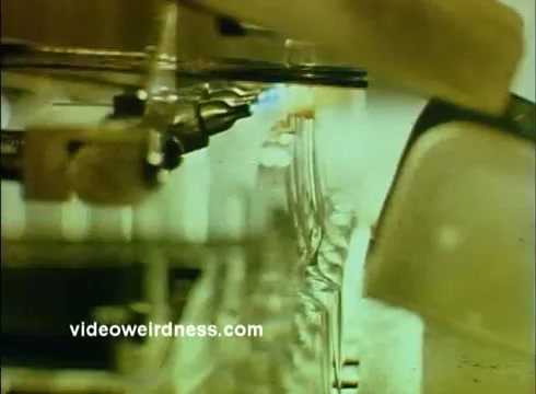 The still from the 'Sandoz Pharmaceuticals/Colors and Chemicals Commercial' shows a close-up of a production line, likely featuring glass vials or containers. There are mechanical elements in the frame, suggesting an industrial or laboratory setting. The overall color palette may lean towards muted greens and yellows typical of 16mm film, emphasizing a scientific atmosphere. The focus on equipment hints at precision and the process of creating pharmaceuticals or chemicals intended for educational use.