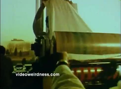 The still from the 'Sandoz Pharmaceuticals/Colors and Chemicals Commercial' depicts a scene where a person is unveiling a large sheet or fabric in front of a machinery, likely related to printing or color processing. The environment appears industrial, with an emphasis on the machinery's role in the production process. The overall color palette suggests a warm, vintage hue typical of 16mm prints, likely aimed at highlighting the technological aspect of Sandoz's contributions to pharmaceuticals and dyes.