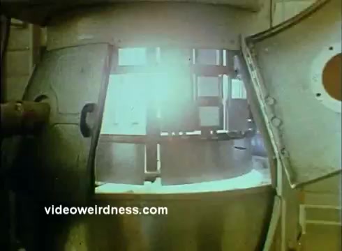 The still from the 'Sandoz Pharmaceuticals/Colors and Chemicals Commercial' depicts a large metallic apparatus, likely a reactor or mixing chamber, with its door partially open. Bright, diffused light emanates from within, suggesting an active process inside. The industrial setting conveys a sense of scientific precision and innovation, aligning with the theme of the commercial aimed at educators in the sciences. The overall visual captures a moment of interest and curiosity in chemical processes.