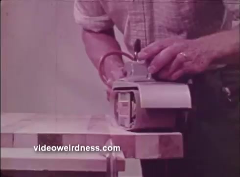 The still features a close-up of a person using a power tool—likely a sander or planer—on a wooden surface. The tool is compact, with a focus on its operational details, suggesting a demonstration of its features. The setting is likely a workshop, emphasizing craftsmanship and the versatility of Black and Decker tools in home improvement projects. The overall tone conveys utility and hands-on engagement with the tools.