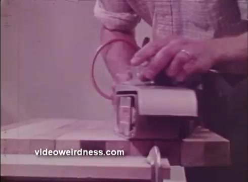 The still from the Black and Decker 1972 promo film features a close-up of a person using a power tool on a piece of wood. The tool appears to be a sander, with a focus on the tool's mechanism and the operator's hand, which shows precision in use. The setting is likely a workshop or home improvement environment, showcasing Black and Decker's range of power tools for DIY projects. The color tone is muted, typical of the era's film quality.