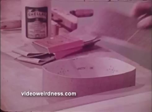The still from the 1972 Black and Decker promo features a workspace with various tools and materials. Prominent items include a can labeled 'Shellac,' a sanding block, and a curved wooden piece, suggesting a focus on woodworking or craft projects. The scene conveys an atmosphere of DIY and tool use typical of home improvement themes in the era.