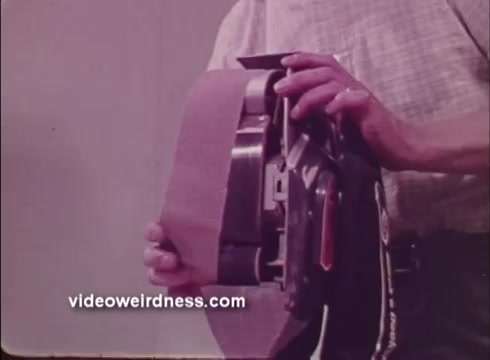 The still features a person holding a power sander, showcasing its sanding belt. The individual appears focused on the tool, indicating a demonstration of its features. The 1972 context suggests a promotional emphasis on the efficiency and design of Black and Decker's power tools. The background is neutral, highlighting the tool's details.