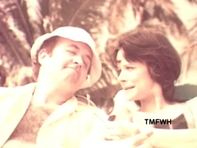 The still from the 'American Express Commercial' featuring Karl Malden captures a relaxed, tropical scene where a man and a woman share a lighthearted moment. The man, wearing a light-colored hat and shirt, has a playful expression, while the woman, dressed in a casual top, appears to be engaged in conversation, possibly laughing. The lush backdrop suggests a vacation setting, reinforcing the theme of travel and leisure. This 16mm print exudes a vintage charm typical of the 1970s commercials, aligning with the tagline 'Don't leave home without them.'